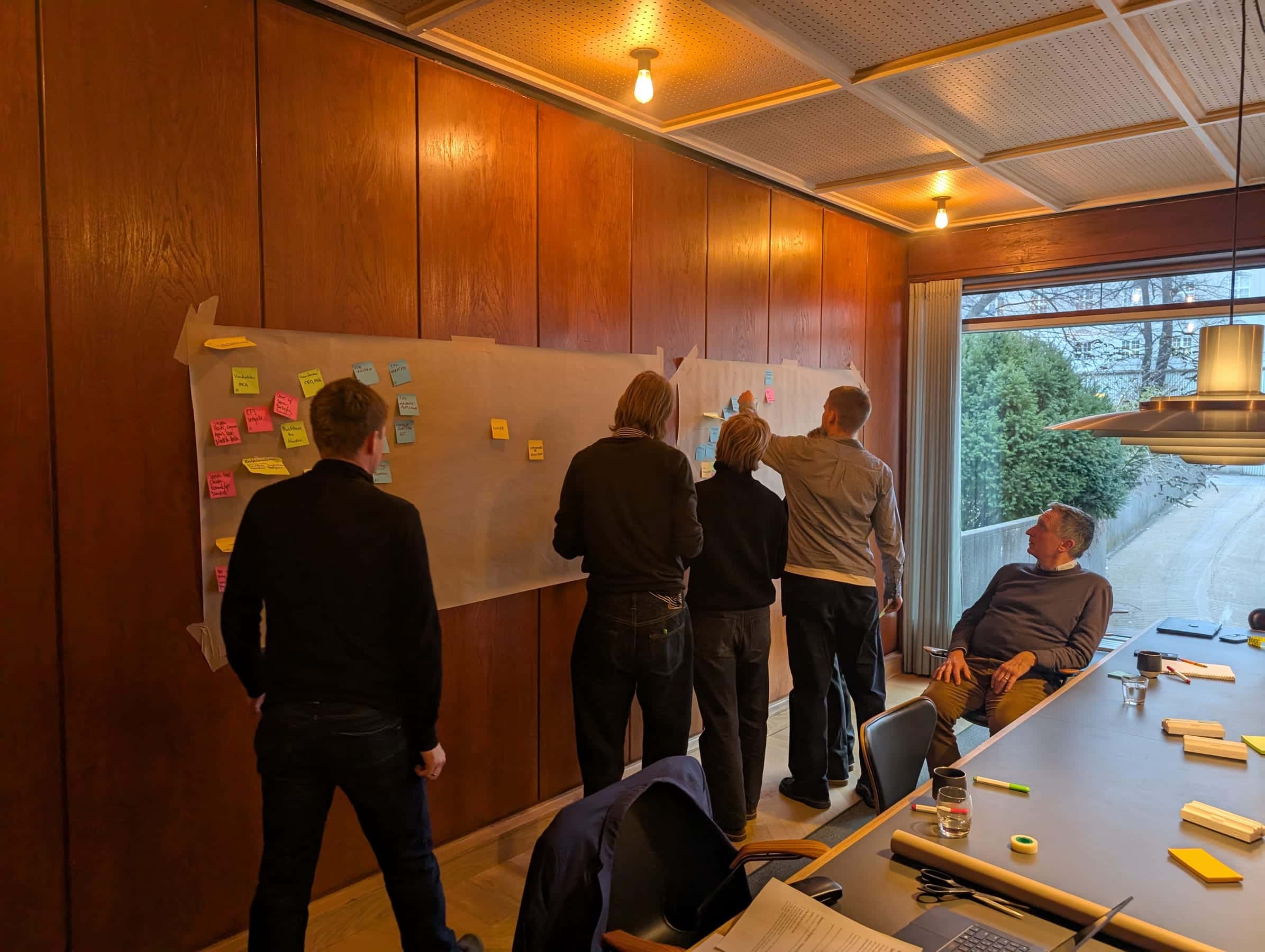 Five people standing at a sticky-note canvas pinned to a wood-panelled wall; one leadership member seated at a long boardroom table observing.