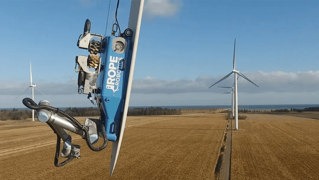 Rope Robotics blue-and-white rig clamped to the leading edge of a wind turbine blade, with another turbine and open farmland visible behind.