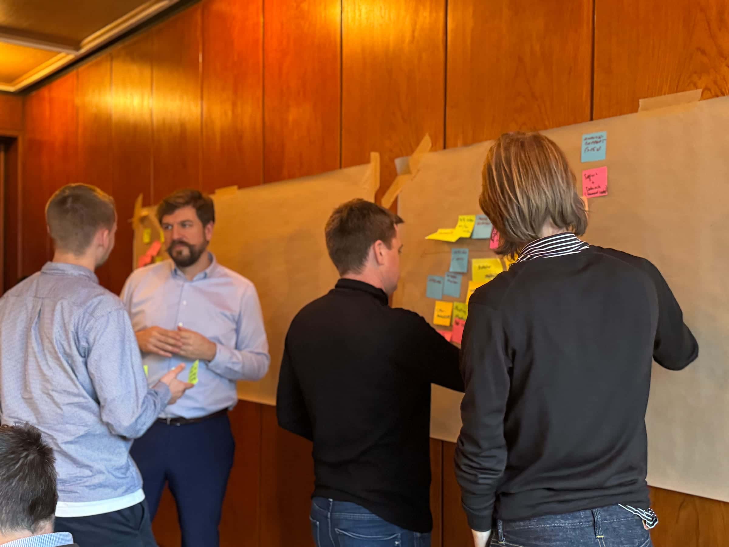 Four people clustered at a brown-paper canvas pinned to a wood-panelled wall, working coloured sticky notes into groups together; one person facilitating the discussion.