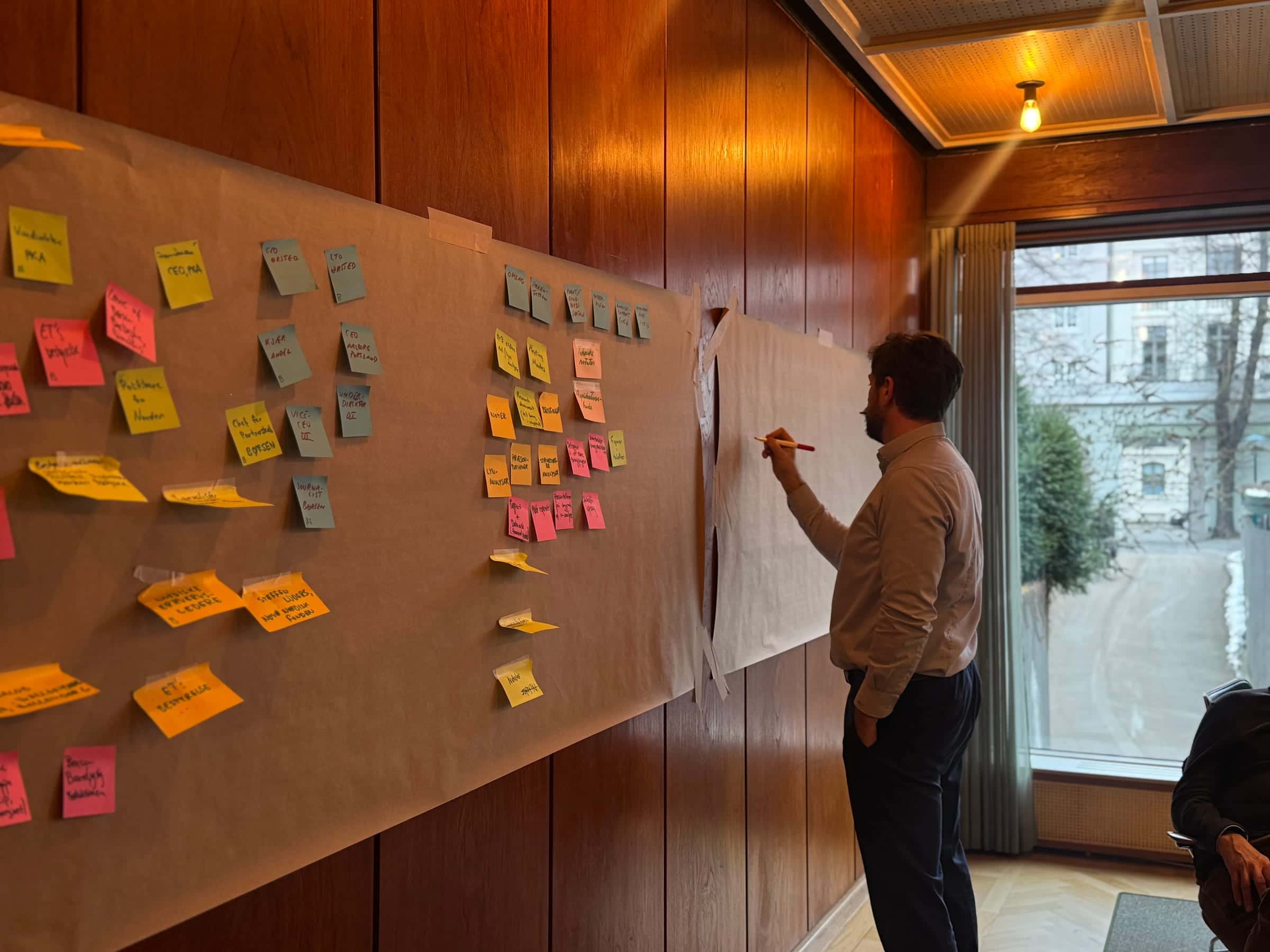 A bearded facilitator in a tan shirt drawing with a red marker on a fresh sheet of brown paper, next to a wall covered in dense clusters of coloured sticky notes — roles, owners and process steps.