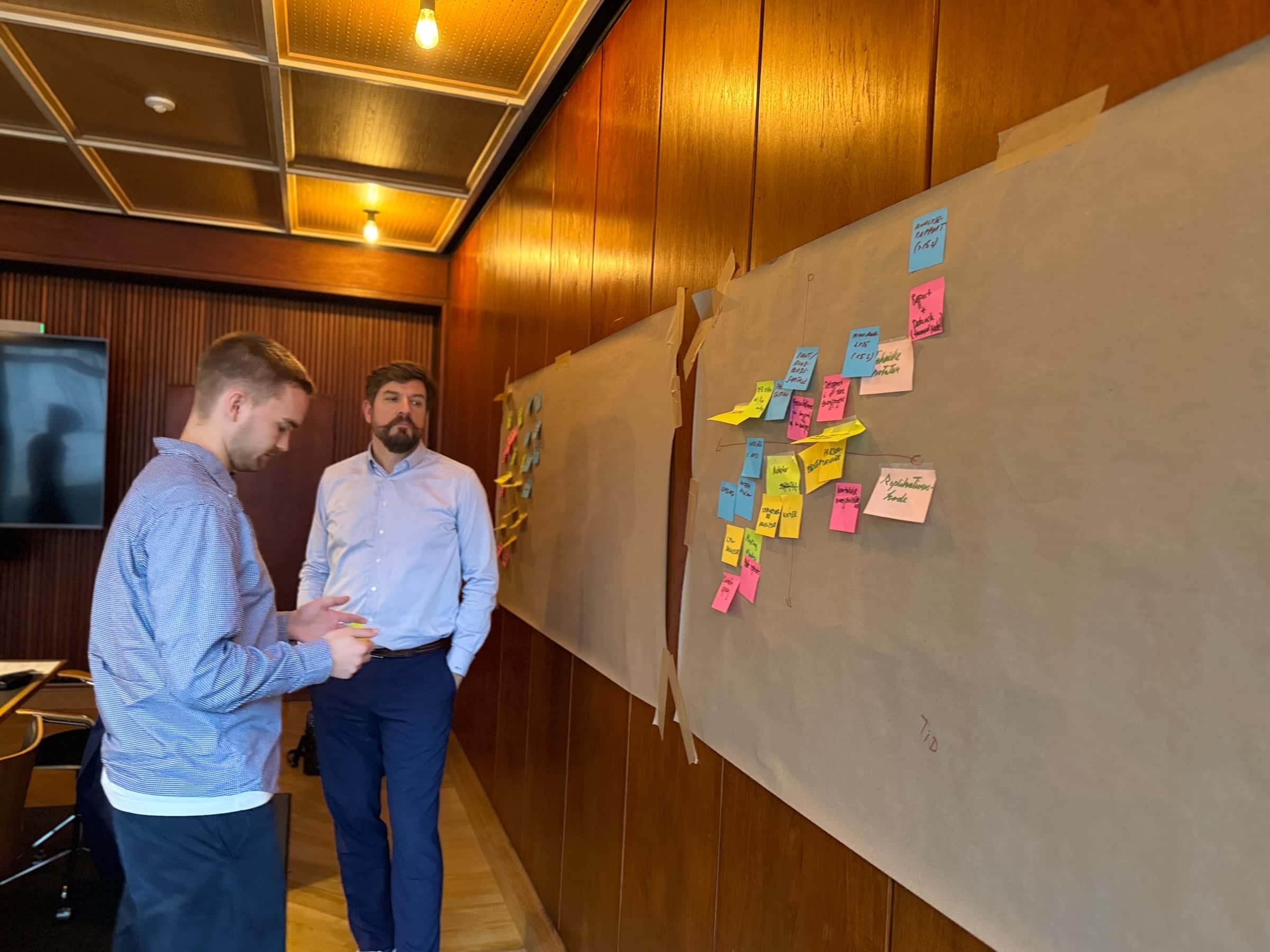Two men in shirts standing and talking quietly side-by-side, with a brown-paper canvas full of coloured sticky notes on the wood-panelled wall behind them.