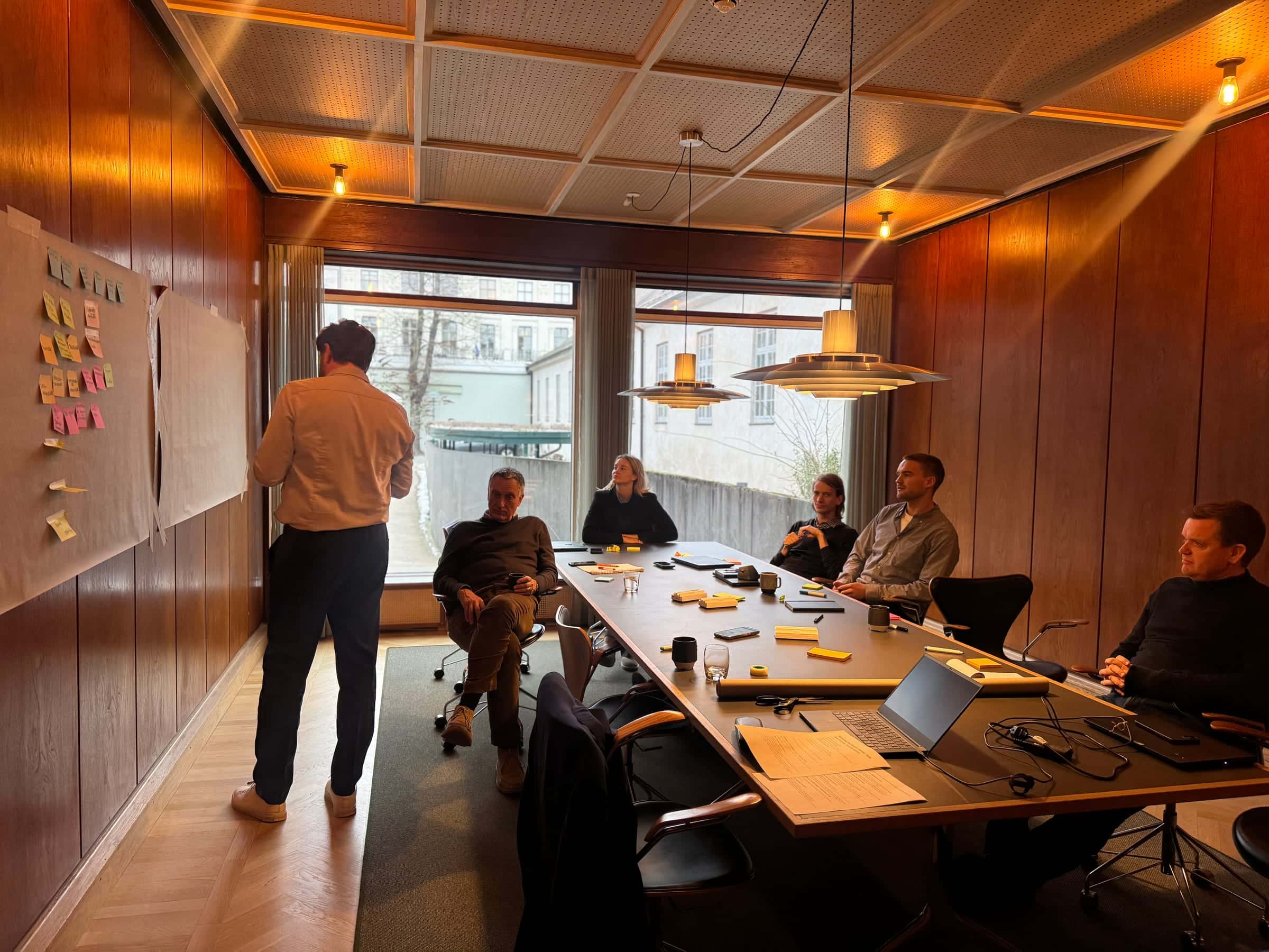 A facilitator stands at a sticky-note canvas at one end of a long boardroom table; five members of a leadership team are seated around the table listening, daylight from tall windows behind them.